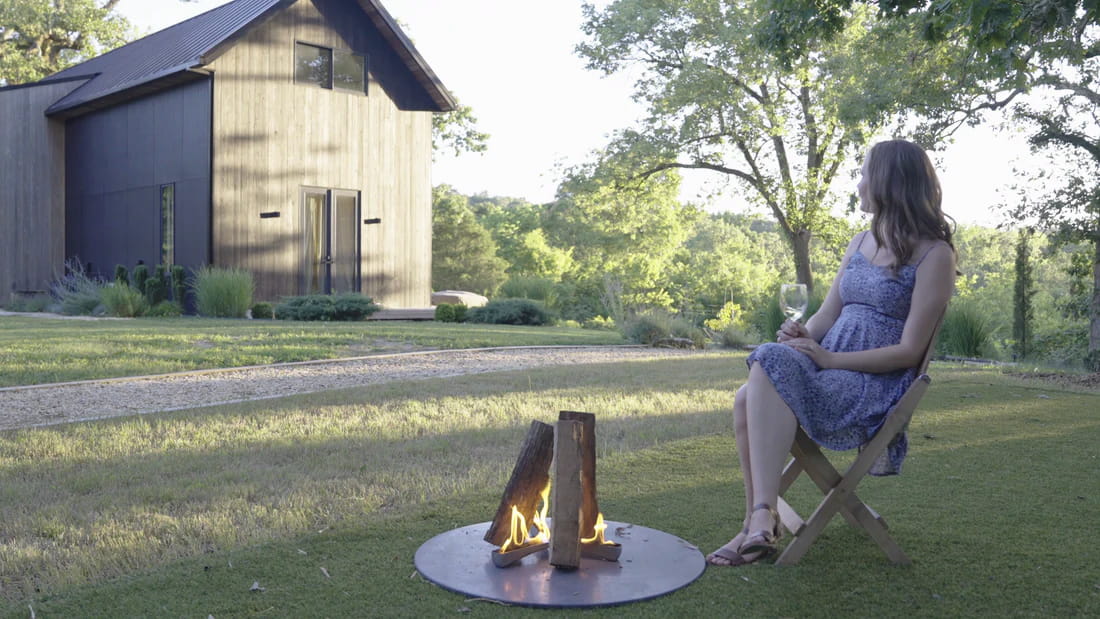 Woman sitting by a fire pit in a backyard with a house and trees in the background